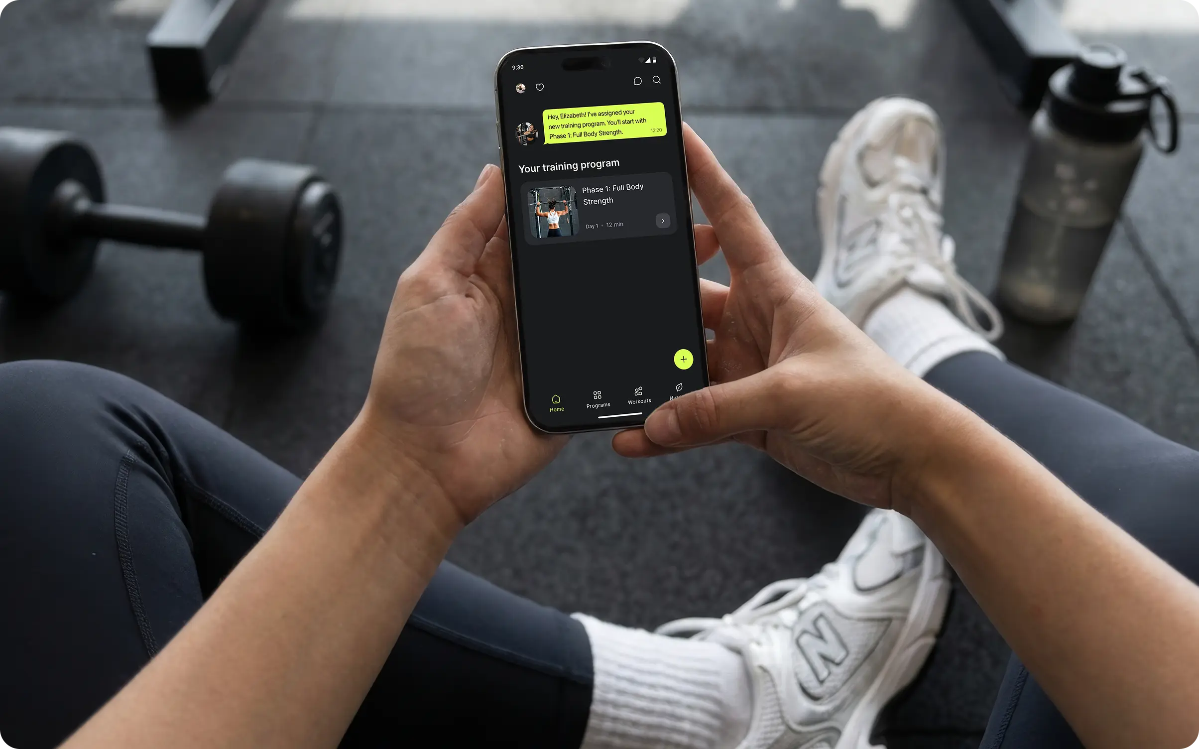 Pessoa em uma academia visualizando o programa de treino atribuído e uma mensagem do treinador no app Ymove no iPhone